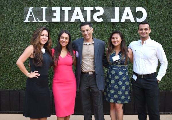 Five smiling alumni posing in front of Cal State LA banner